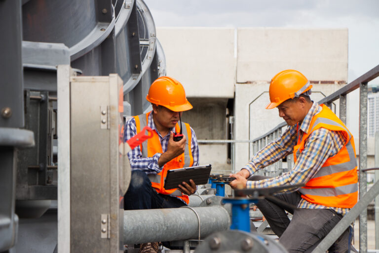 Asian male engineer inspecting pipe fittings and chillers, refrigeration plant refrigeration plant in factory. male mechanic wearing helmet and Reflective Safety Vest uses laptop, tablet, equipment with sets of cooling towers in conditioning systems back